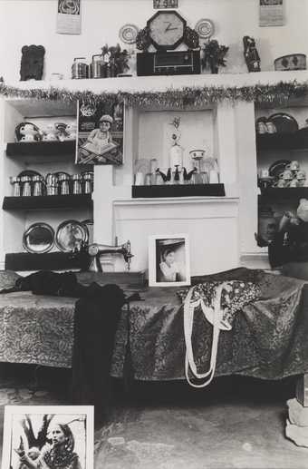 An interior scene showing a bed, framed photographs, a garment and a bag, with various ornaments and a clock in the background.