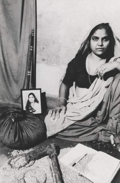 A figure sits on the floor in an indoor setting, surrounded by a cloth bag, an axe, a staff, a notebook and pen, some jewellery, a photograph and some loose grain.