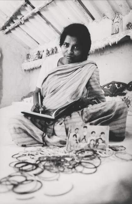 A figure sits cross-legged on the floor in an indoor setting, writing in a notebook and surrounded by a photograph of a group of figures and some jewellery.