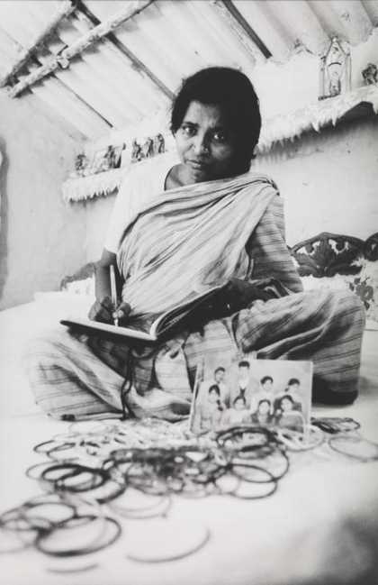 A figure sits cross-legged on the floor in an indoor setting, writing in a notebook and surrounded by a photograph of a group of figures and some jewellery.