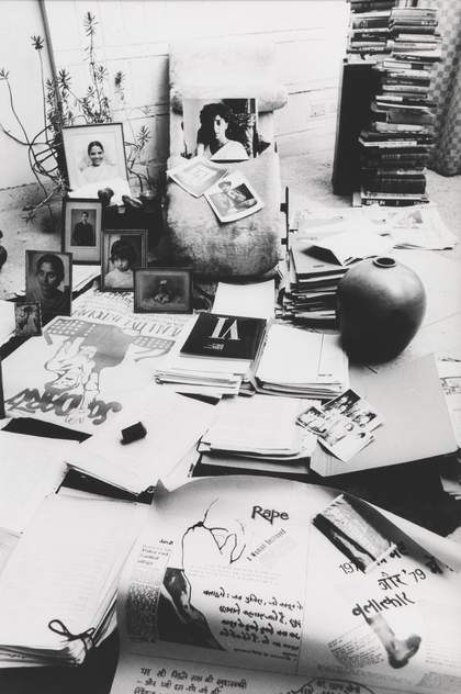An interior scene showing posters, written texts, a stack of books, photographs and a vase.