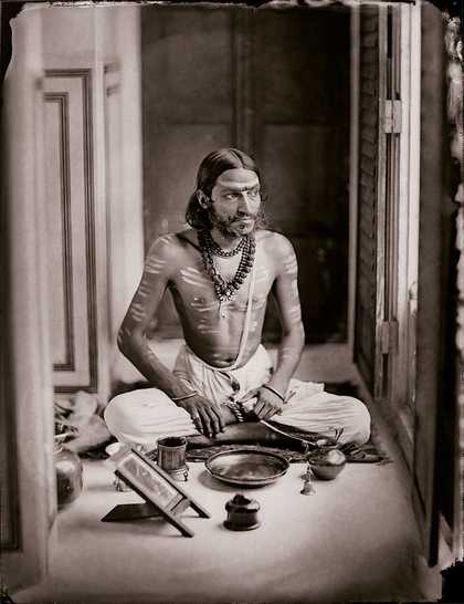 A figure sits cross-legged on a set of striped cushions, wearing a sash and holding a shield-like object, with an ornately decorated floral backdrop and a sun-like motif behind them.