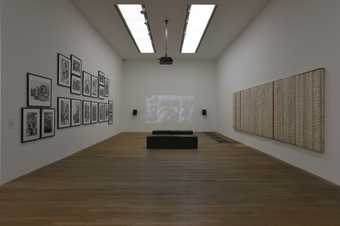 A gallery with a set of framed photographs hanging on the left wall, a film projected in the middle and a long, horizontal textile artwork handing on the right wall.