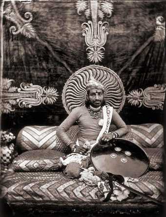 A figure sits cross-legged on the floor in an indoor setting surrounded by a bowl, a mirror and some small pots, with beads around their neck and pale horizontal lines painted on their arms and torso.