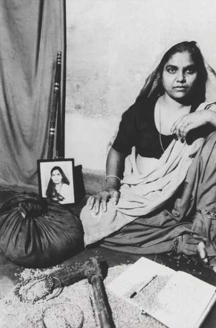 A figure sits on the floor in an indoor setting, surrounded by a cloth bag, an axe, a staff, a notebook and pen, some jewellery, a photograph and some loose grain.
