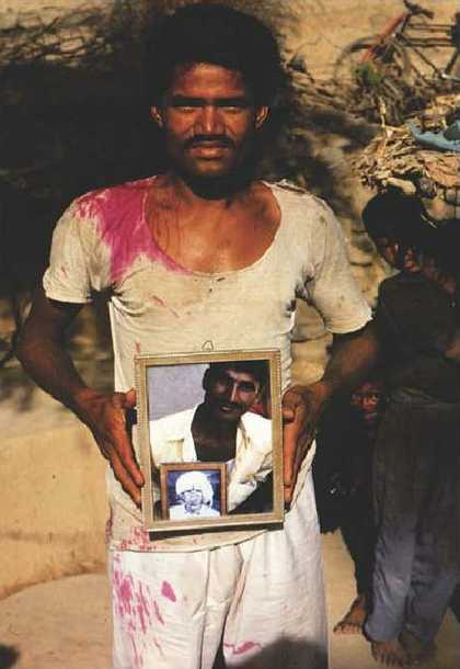 A figure stands in an outdoor setting holding up a framed photograph that features a figure holding up a photograph of another figure.