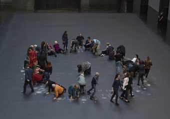 A group of people in a circle on the dark grey ground of the Turbine Hall, standing, sitting, crouching - where their bodies have touched the ground they've left white marks