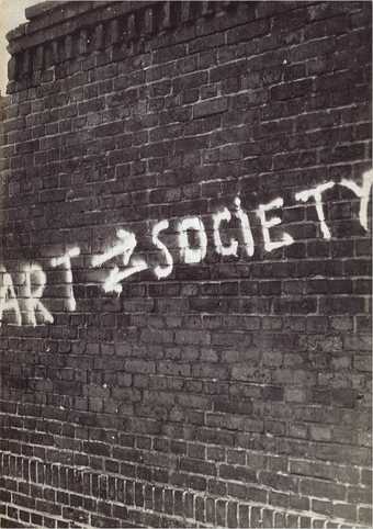 Black-and-white photograph of a brick wall with white graffiti on it reading ART SOCIETY with two arrows between the two words, the upper pointing right (towards SOCIETY), the lower pointing left (towards ART) 