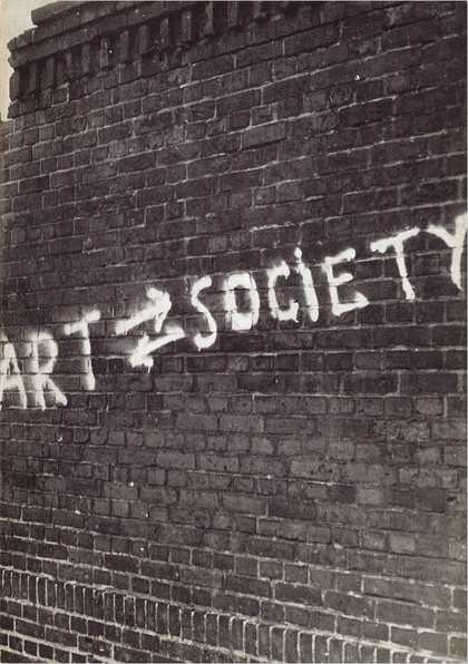 Black-and-white photograph of a brick wall with white graffiti on it reading ART SOCIETY with two arrows between the two words, the upper pointing right (towards SOCIETY), the lower pointing left (towards ART)