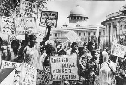 A large crowd of figures march and hold up banners in an outdoor setting.