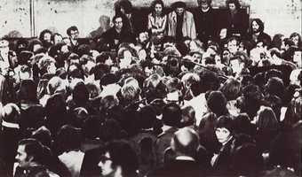 Black-and-white photograph of a tightly packed crowd of people, attention focused on speakers in the centre of the group