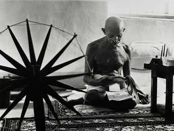 A figure sits cross-legged on the floor in an indoor setting, reading some papers and surrounded by an ornate rug and a spinning wheel.