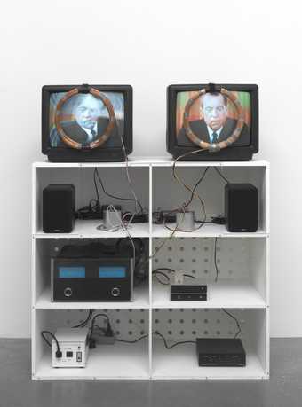 Photograph of an installation composed of two television monitors, both showing footage of Richard Nixon; each monitor is positioned on top of a white shelving unit, placed side by side, with a video switcher and audio system on the shelves below