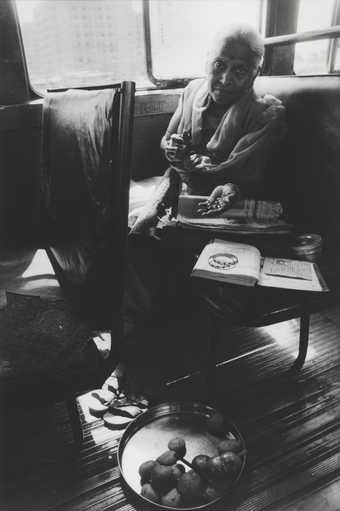 A figure sits in an interior setting holding a set of marbles, with a book, jewellery and a tin containing large pebbles in front of them.