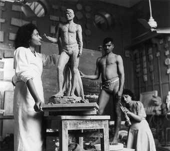 An artist’s studio setting in which a figure stands looking up at a sculpture of a male body, while another figure crouches down next to a live model.