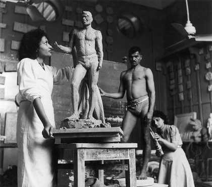 An artist’s studio setting in which a figure stands looking up at a sculpture of a male body, while another figure crouches down next to a live model.