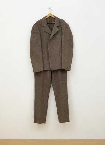 Photograph of a two-piece suit comprising a jacket and a pair of trousers made from coarse grey felt, on a clothes hanger, here shown hanging from a wooden coat hanger on a white gallery wall.