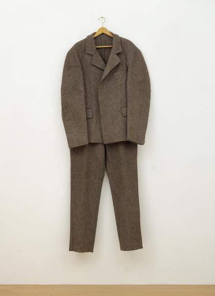 Photograph of a two-piece suit comprising a jacket and a pair of trousers made from coarse grey felt, on a clothes hanger, here shown hanging from a wooden coat hanger on a white gallery wall.