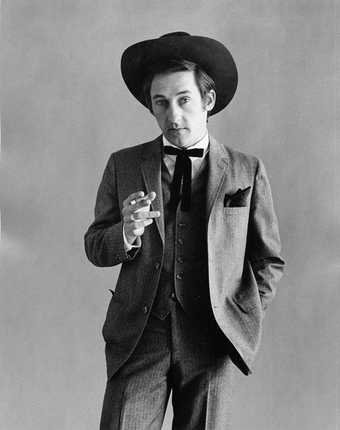 Fig.3 Black-and-white studio portrait photograph of Ed Ruscha dressed as a cowboy, left hand in his pocket, right hand holding a cigarette like a gun pointed to the camera