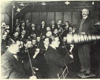 A black and white photograph of a figure standing in front of an orchestra and next to a large, cone-shaped horn