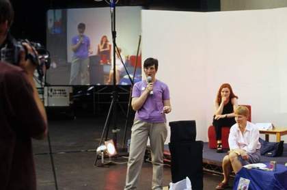 A person stands in front of a white backdrop and speaks into a microphone while being filmed, with an image of the filmed scene projected onto a large screen in the background.