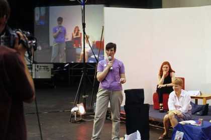 A person stands in front of a white backdrop and speaks into a microphone while being filmed, with an image of the filmed scene projected onto a large screen in the background.