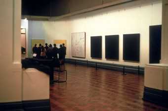 Photograph of an art gallery with abstract paintings hanging on the walls; a group of people are gathered around viewing one of the paintings