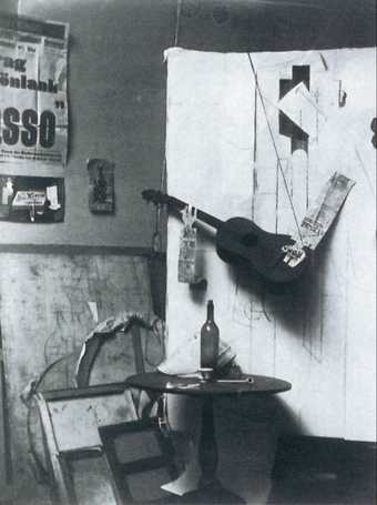 A large white sheet with abstract lines drawn onto it, with a guitar and newspaper hanging down and resting on its surface and a table and wine bottle positioned in front of it