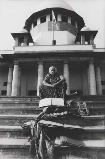 A figure sits at the top of some steps leading up to a large building with columns, with documents, a framed portrait and swathes of cloth placed on the steps in front of them.