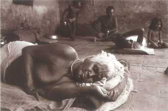 A figure lies with eyes closed on a mat on a flagstone floor, with other figures sitting and lying in the background.