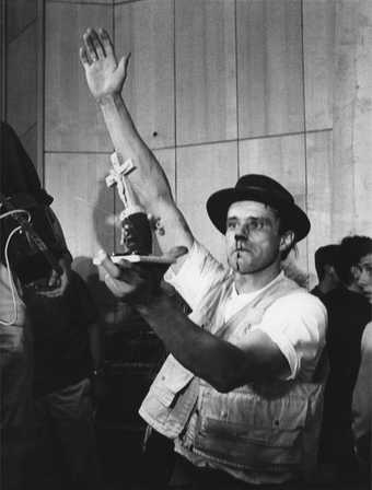 Black-and-white photograph of Joseph Beuys, blood dripping from his nose, right arm raised, left hand holding up a small sculptural object featuring a crucifix