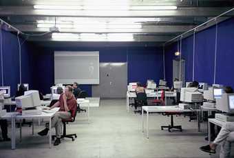 A room featuring several desks and a number of computers, with individuals sitting at them and using them.