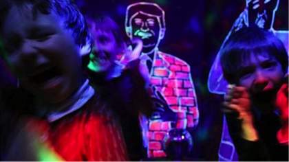 Children smiling and shouting in a dark space against a backdrop of neon-painted figures.