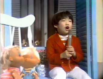 Video still showing a young boy wearing a red cardigan, singing into a microphone, with two dolls in the foreground