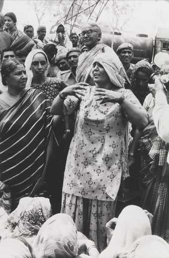 A figure speaks and gesticulates to a large crowd of people in an outdoor setting.