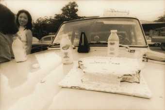 Black-and-white photograph of a car bonnet upon which sit a rectangular cake iced with the words ‘lemon cake’, with sections eaten from two corners, and three bottles of chocolate milk and a metronome. Two women stand next to the car.