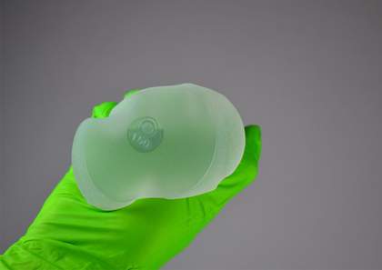 A semi-transparent breast implant held by a hand in a green plastic glove.