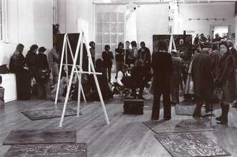 Black-and-white photograph of a studio with easels and blackboard panels. Joseph Beuys is at the centre while a crowd of people watch him