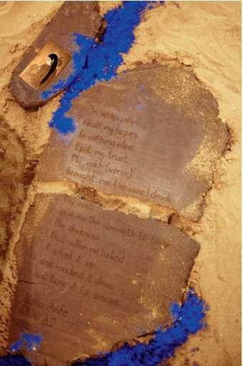 A stone tablet with writing carved into it, surrounded by patches of sand and dry pigment in yellow and blue.