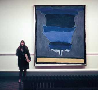 Photograph of Helen Frankenthaler’s 1964 painting Cape, (Provincetown) hanging on a gallery wall with a woman standing next to it; the painting is abstract in a range of blues with a sandy yellow horizontal strip at the bottom