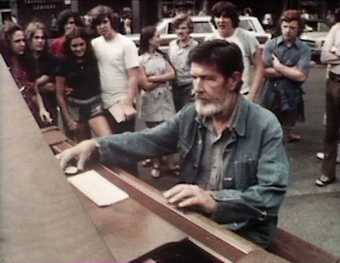Video still showing John Cage sitting at a piano outdoors in Harvard Square, surrounded by people watching the performance