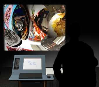 An individual stands next to a computer screen and in front of a colourful, distorted image that is projected on the wall.