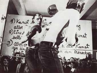 Black-and-white photograph of two men boxing in a ring in the foreground, with a crowd watching. Behind the audience is a large white panel with three statements written on it in German