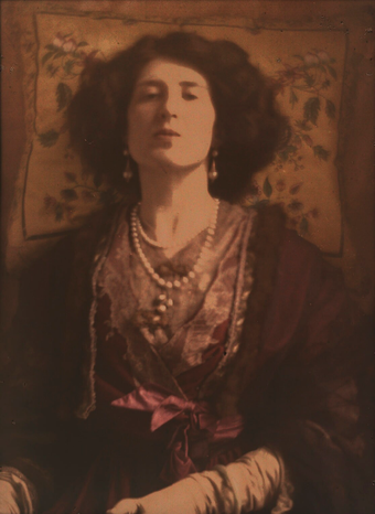 The head and torso of a figure faces the viewer, with the head tilted backwards, a flowered cushion behind the head and strings of pearls around the neck
