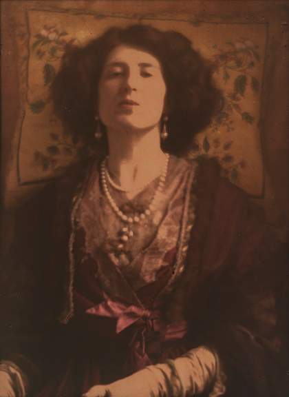 The head and torso of a figure faces the viewer, with the head tilted backwards, a flowered cushion behind the head and strings of pearls around the neck