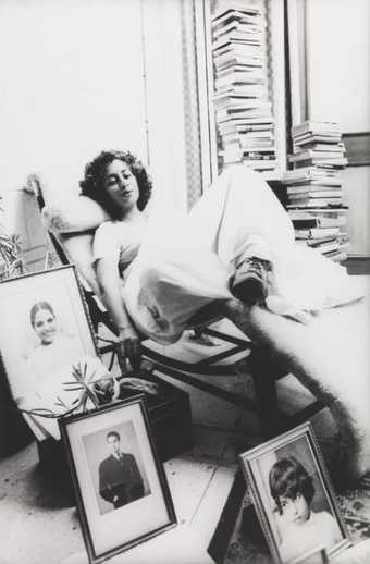 A figure lies on a reclining chair in an indoor setting, surrounded by framed portrait photographs and stacks of books.