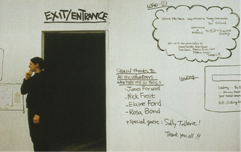 A person stands in front of a white wall and next to a dark doorway that is marked ‘Exit/Entrance’. Various pieces of text are hand-written on the wall, including exhibition acknowledgements.