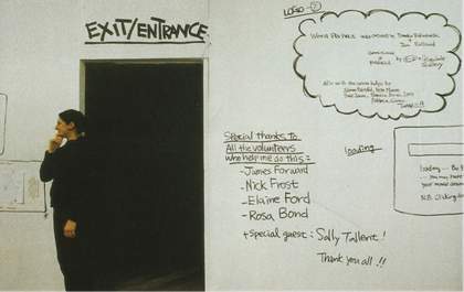 A person stands in front of a white wall and next to a dark doorway that is marked ‘Exit/Entrance’. Various pieces of text are hand-written on the wall, including exhibition acknowledgements.