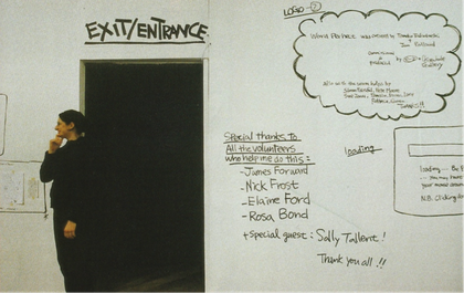 A person stands in front of a white wall and next to a dark doorway that is marked ‘Exit/Entrance’. Various pieces of text are hand-written on the wall, including exhibition acknowledgements.