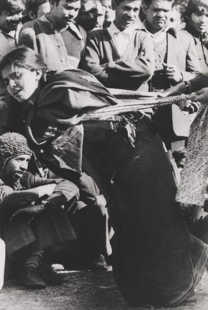 A figure leans forward, pushing against a band of cloth that is wrapped around them and held by another figure, while a crowd looks on.